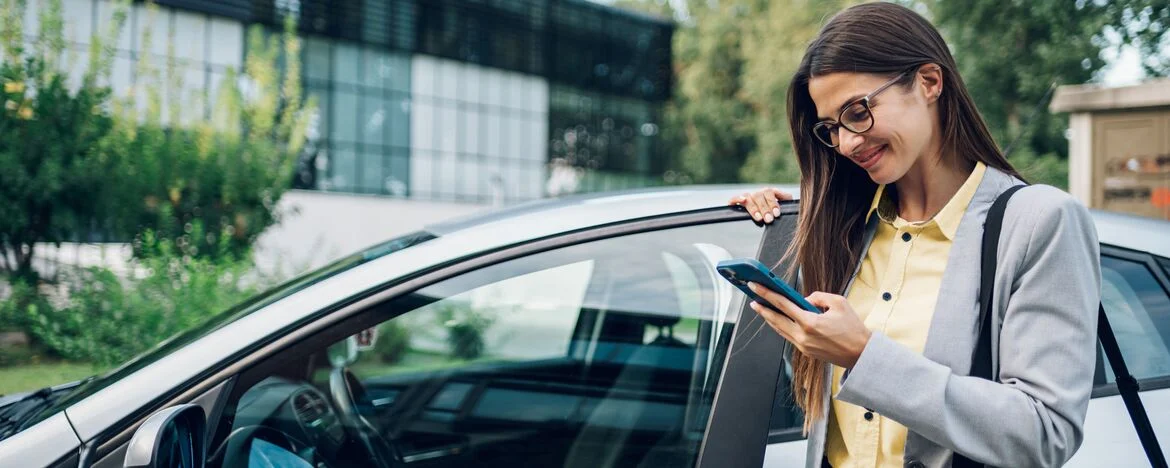 Woman dressed in grey suit and yellow shirt getting out of her car and looking at her phone