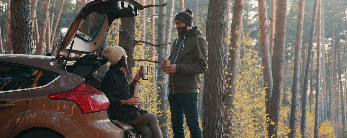 Person sat in back of their car drinking a warm drink and talking to a friend