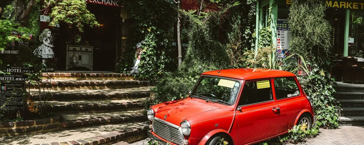 An old, red car in an abandoned city and surrounded by overgrown foliage