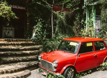 An old, red car in an abandoned city and surrounded by overgrown foliage